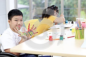 Disabled kids classroom, happy smiling schoolboy on wheelchair showing painted hands, children have fun together during study at