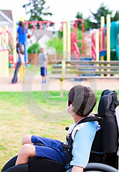 Disabled little boy in wheelchair watching children play on play