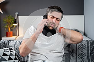Disabled man with neck brace talking on phone with doctor, experiencing pain while sitting on couch in the living room