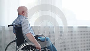 Disabled old man is sitting in a wheelchair at home alone. A handicapped person in a wheelchair is looking out the