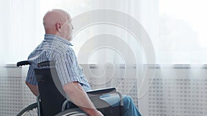 Disabled old man is sitting in a wheelchair at home alone. A handicapped person in a wheelchair is looking out the