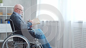 Disabled old man is sitting in a wheelchair at home alone. A handicapped person in a wheelchair is reading the book