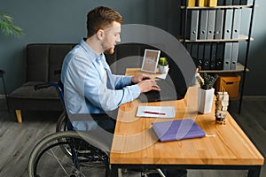 Disabled person in the wheelchair works in the office at the computer. He is smiling and passionate about the workflow