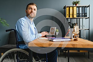 Disabled person in the wheelchair works in the office at the computer. He is smiling and passionate about the workflow.