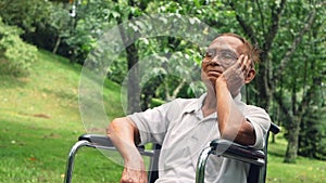 Disabled senior man in eyeglasses sitting in wheelchair looking away and thinking with his hand on chin in the park.