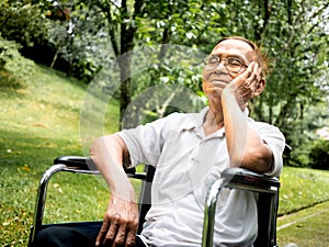 Disabled senior man in eyeglasses sitting in wheelchair looking away and thinking with his hand on chin in the park.