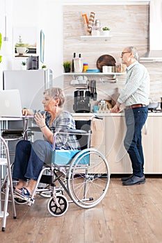 Disabled senior woman in wheelchair working from home at laptop in kitchen