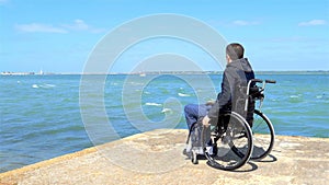 Disabled young man sitting in a wheelchair and looks at the sea