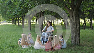 Disabled young teacher reading book to boys and girls in park