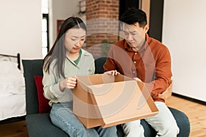 Disappointed asian married spouses unpacking cardboard box, received damaged item or unwanted stuff, sitting on sofa