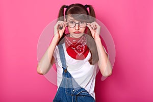 Disappointed charismatic young lady with bangs and long pigtails has frown face, touches her eyeglasses, choosing comfortable