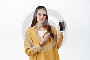 Disappointed female customer pointing at smartphone screen and frowning upset, complaining on bad delivery shopping app