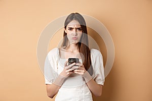 Disappointed girl with smartphone frowning, pucker lips upset, reading bad news on phone, standing on beige background