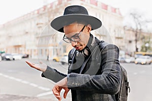 Disappointed man looking at wristwatch while waiting someone. Worried african guy in hat standing on the street..