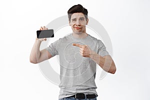 Disappointed man pointing at lame online content, showing empty smartphone screen and frowning displeased, show dislike