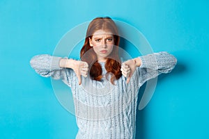 Disappointed redhead girl in sweater showing thumbs-down, judging bad product, disagree and dislike promo, standing over