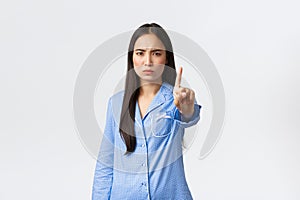 Disappointed serious and angry asian woman in blue pajamas shaking finger and frowning displeased, forbid something