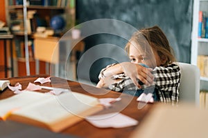 Disappointed upset primary child school girl sitting alone hugging knees in front of desk with difficult homework.