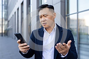 A disappointed young Asian businessman spreads his hands while looking at the mobile phone screen, standing outside an