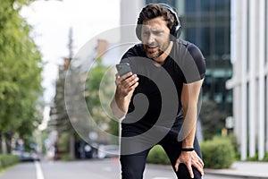 Disappointed young man sportsman runner standing bent and tired on the street wearing headphones and looking upset at
