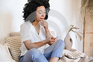 Disappointing afro american woman sitting on bed, holding pregnancy test