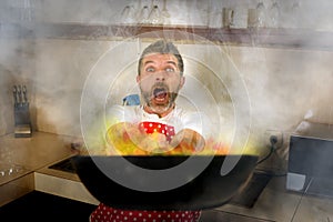 Disaster home cook at kitchen- young funny and desperate man in cooking apron holding pan in flames in stress and fear making a