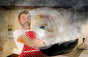 Disaster home cook at kitchen- young funny and desperate man in cooking apron holding pan in flames in stress and fear making a