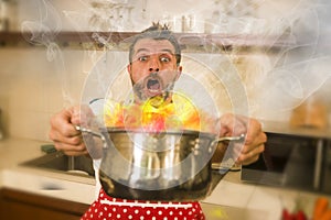 Disaster home cook at kitchen- young funny and desperate man in cooking apron holding pot in flames in stress and fear making a