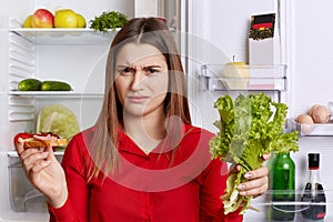 Discontent female frown face, has to keep to diet, eats only fruits and vegetables, holds lettuce and sandwhich in hands, stands a