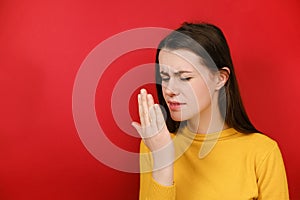 Discontent young brunette woman checking her breath with hand, smells something awful, pinches nose, frowns in displeasure