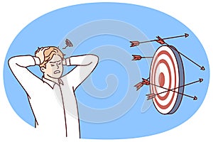 Discouraged man clutching head near darts board after failed attempts to hit target. Vector image