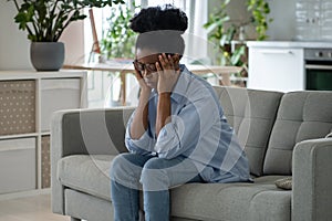 Discouraged sad African American woman sits on sofa and feeling stressed because of loneliness