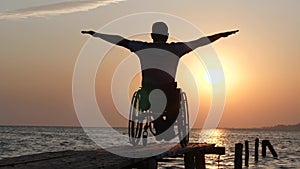 Diseased man in wheelchair relaxes and lift his hands backdrop of orange sky on summer trip