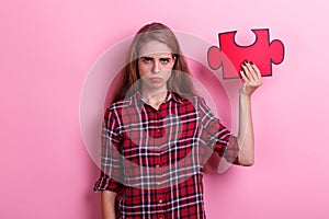 Disgruntled girl, holds a red puzzle on her raised hand. On a pink background.