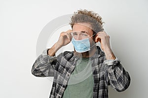 Disgustedly under duress puts on protective medical mask on his face to protect coronavirus, with curly hair, young man