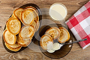 Dish with pancakes, small pancakes poured condensed milk, spoon in saucer, bowl with condensed milk, napkin on wooden table. Top