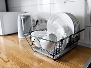 Dish rack holds many dishes and cups against wooden countertop, white wall tiles, sink and faucet. Budget and lightweight