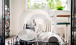 Dish washing machine. Inside an open dishwasher, view to kitchen. Clean plates, utensils, cutlery and pot in washer rack.