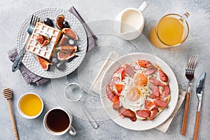 Dishes for home cooked hearty Breakfast. Fried eggs with sausages and tomatoes. Belgian fluted waffles with figs and grapes. Gray