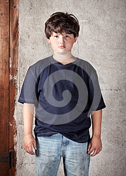 Disheveled and Dirty Caucasian Boy in Jeans and Blue T-Shirt Standing In Front of Stucco Wall