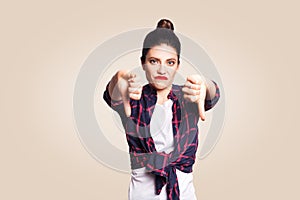 Dislike. Young unhappy upset girl with casual style and bun hair thumbs down her finger, on beige blank wall with copy space looki