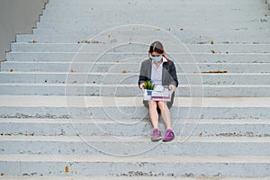 Dismissed woman with a box of personal belongings sits on the stairs on the outside. Female employee in medical mask
