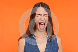 Disobedient crazy brunette woman in denim clothes sticking out tongue and grimacing with closed eyes