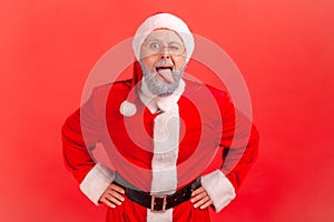 Disobedient funny elderly man with gray beard wearing santa claus costume demonstrating tongue, behaving naughty unruly, childish