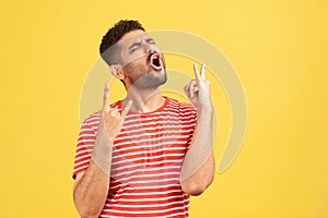 Disobedient naughty man with beard in striped t-shirt showing rock and roll gesture and excitedly screaming, having fun, enjoying