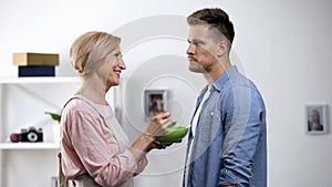 Displeased adult man refusing mother feeding from spoon, overprotection problem