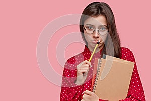 Displeased attractive young lady writes records in notepad, carries spiral notebook, has round big spectacles, models