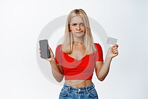 Displeased blond girl grimaces, shows mobile phone screen and credit card, pouting upset, standing over white background