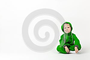 A displeased charismatic child in a bright green overalls sits on the floor and looks at the camera with doubt and contempt