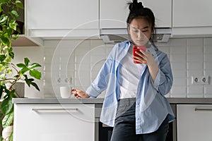 Displeased embarrassed Asian woman with phone standing in kitchen reads bad notification from bank